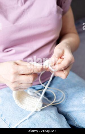 Hände einer erwachsenen Frau, die einen weißen Schal mit Stricknadeln strickt, während sie auf dem Sofa sitzt Stockfoto