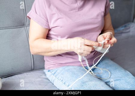 Hände einer erwachsenen Frau, die einen weißen Schal mit Stricknadeln strickt, während sie auf dem Sofa sitzt Stockfoto