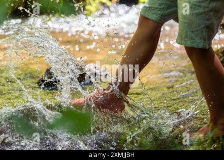 Kind in Shorts, das barfuß in einem Fluss frisches Wasser spritzt. Planschspiel, indem Sie Flusswasser treten. Nahaufnahme der Füße im Fluss. Stockfoto