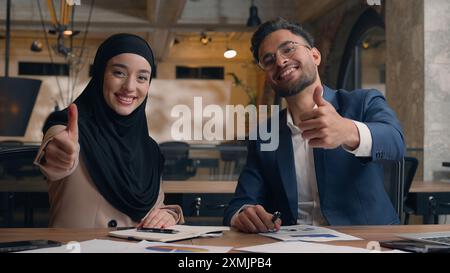 Zwei Geschäftspartnerkollegen glückliche männliche Kollegen, die im Büro schreiben arabischer indischer Mann muslimische Frau im Hijab zeigen Daumen nach oben Geste gut Stockfoto