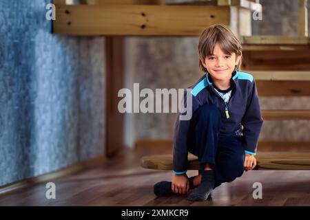 Ein lächelndes, schlankes europäisches männliches Kind in Sportuniform sitzt auf der Stufe einer Holztreppe, die zum zweiten Stock des Landhauses führt. Stockfoto