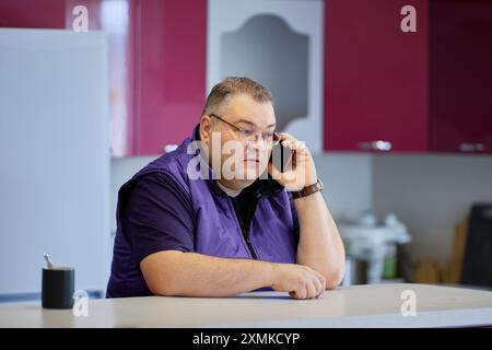 Ernsthaftes Gespräch am Telefon, großer Kaukasier in seinen 40ern mit Brille und kurzen Haaren benutzt Smartphone in der Küche. Stockfoto