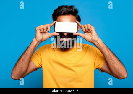 Lächelnder Mann, Der Das Smartphone Vor Dem Gesicht Gegen Blau Hält Stockfoto