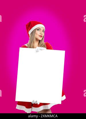 Wunderschöne junge lateinfrau, die als Weihnachtsmann verkleidet ist und in die Kamera blickt und ein Schild als Nachahmung auf magentafarbenem Hintergrund zeigt Stockfoto
