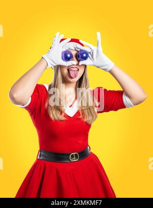 Junge Frau Weihnachtsmann mit zwei lila weihnachtskugeln in den Augen, die ihre Zunge auf gelbem Hintergrund zu weihnachten ausstreckt Stockfoto