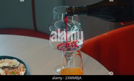 Person, die Rotwein in Glas an einem gemütlichen Restauranttisch mit Essen gießt, Stockfoto