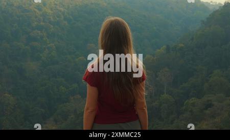 Eine Frau mit langen Haaren steht auf einer Klippe mit Blick auf den üppigen, grünen Wald Stockfoto