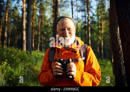 Mittleres Porträt eines fröhlichen männlichen Seniortouristen mit Bart auf dem Gesicht, das Fernglas hält und in die Kamera blickt, während er im Wald steht Stockfoto