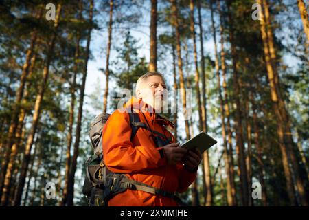 Niedriger Blickwinkel auf ältere kaukasische männliche Touristen mit orangefarbener Jacke mit digitalem Tablet während der Wanderung am Sommertag Stockfoto