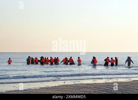 Eine Gruppe von Menschen, die als Migranten gelten, waten durch das Meer, um sich auf ein kleines Boot vor dem Strand in Gravelines, Frankreich, vorzubereiten. Bilddatum: Montag, 29. Juli 2024. Stockfoto