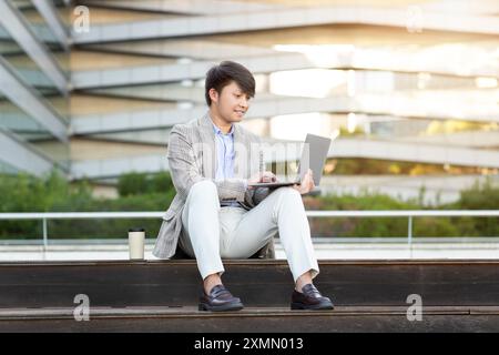 Junger Mann, der vor dem modernen Gebäude an einem Laptop arbeitet Stockfoto