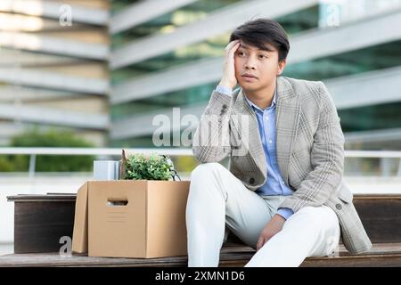 Deprimierter junger Mann, der auf der Bank sitzt, mit einer Schachtel voller Sachen Stockfoto