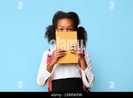 Schwarze Schülerin versteckt sich hinter Buch über gelben Hintergrund Stockfoto