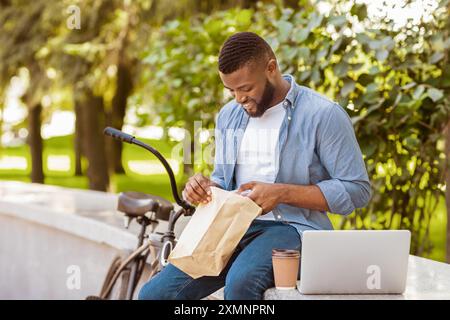 Mittagessen Im Park. Black Guy, Der Im Freien Mit Laptop Isst Und Ausruht Stockfoto