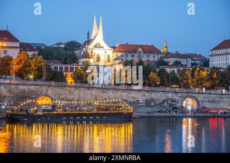 Die Moldau fließt am beleuchteten Emmaus-Kloster in Prag, Tschechien, vorbei. Das Wasser reflektiert die Lichter der Stadt und schafft eine wunderschöne Szene. Ein Boot liegt nahe dem Fluss an. Stockfoto