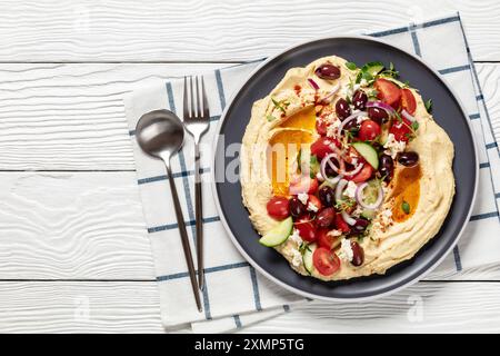 Griechischer Hummus mit frischen Gurken, Oliven, Tomaten, Fetakäse, roten Zwiebeln und Thymian auf Teller auf weißem Holztisch mit Löffel und Gabel, Hori Stockfoto