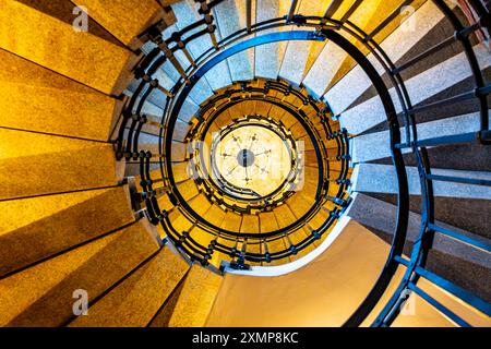 Ein Blick vom Boden einer Wendeltreppe, mit Blick auf einen Kronleuchter. Die Treppe verfügt über einen hölzernen Handlauf und Metallgeländer. Die umliegenden Wände sind in warmer, blassgelber Farbe. Stockfoto
