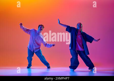 Dynamisches Bild von jungen Mann und Frau in lässiger übergroßer Kleidung, die Hip-Hop vor verlaufendem Studiohintergrund im Neonlicht tanzen Stockfoto