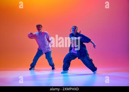 Dynamisches Bild von jungen Mann und Frau in lässiger übergroßer Kleidung, die Hip-Hop vor verlaufendem Studiohintergrund im Neonlicht tanzen Stockfoto