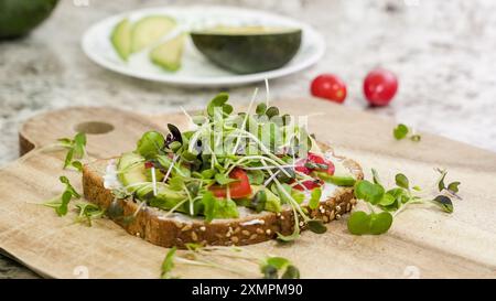 Ein köstliches Sandwich mit offenem Gesicht wird auf einem Holzbrett mit einer großzügigen Schicht Avocado, geschnittenen Tomaten und einer Fülle von lebhaften mi präsentiert Stockfoto