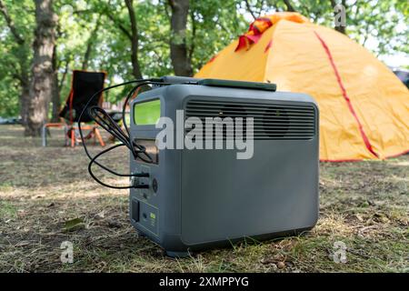 Tragbares Kraftwerk mit Lithium-Ferrophosphat-Batterie, das elektronische Geräte auflädt, die auf einem wilden Campingplatz vor dem gelben Zelt verwendet werden. Tragbar Stockfoto