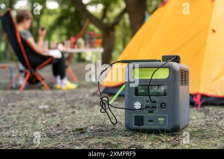 Tragbares Kraftwerk mit Lithium-Ferrophosphat-Batterie, das elektronische Geräte auflädt, die auf einem wilden Campingplatz vor dem gelben Zelt verwendet werden. Tragbar Stockfoto
