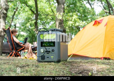 Tragbares Kraftwerk mit Lithium-Ferrophosphat-Batterie, das elektronische Geräte auflädt, die auf einem wilden Campingplatz vor dem gelben Zelt verwendet werden. Tragbar Stockfoto