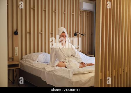 Frau in weißem Bademantel, die auf dem Bett mit Holztäfelung sitzt. Moderne Inneneinrichtung für Wohnkultur und Entspannung. Stockfoto