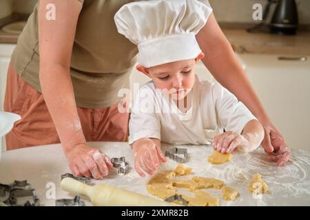 Eine Frau hilft einem kleinen Kind, Kekse zu machen. Das Kind trägt einen Kochhut und verwendet Keksschneider, um den Teig zu Formen. Die Szene ist warm Stockfoto