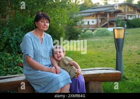 Frau und Kind sitzen auf Holzbank in einem Garten mit Haus im Hintergrund. Familienporträt. Kind im Alter von drei Jahren (dreijähriger Junge) Stockfoto