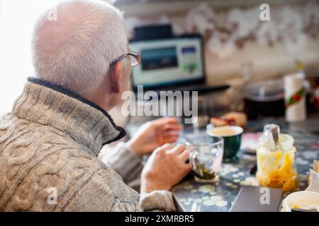 Ein älterer Mann sitzt an einem Küchentisch und genießt seinen Morgenkaffee. Er konzentriert sich auf einen Laptop, der Informationen anzeigt, während er von verschiedenen Snacks umgeben ist Stockfoto