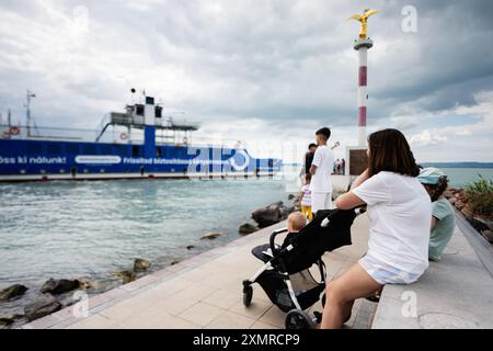 Eine Familie, die mit einem Kind im Kinderwagen am Wasser sitzt, beobachtet an einem bewölkten Tag eine große blaue Fähre Stockfoto