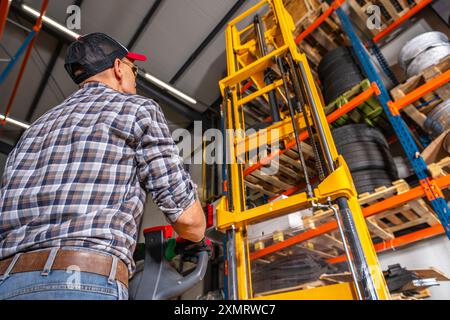 Ein Lagerarbeiter betreibt einen Gabelstapler, um Reifen in Regalen in einem Lagerbereich abzuholen. Der Arbeiter ist konzentriert und trägt Sicherheitsausrüstung, Surround Stockfoto