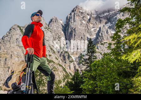 Ein Wanderer in leuchtend roter Jacke und Mütze steht selbstbewusst mit Trekkingstöcken und blickt auf die majestätischen Berge. Üppig grüne Bäume umgeben die Szene, A Stockfoto