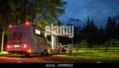 Ein Freizeitfahrzeug parkt auf einem von warmen Lichtern beleuchteten Campingplatz, umgeben von hohen Bäumen und Bergen. Eine Person entspannt sich draußen auf einem Stuhl Stockfoto