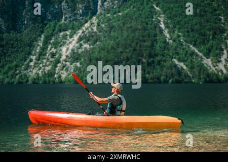 Eine Person Kajak auf einem ruhigen See umgeben von steilen, üppigen Bergen unter einem klaren blauen Himmel. Das leuchtend orange und gelbe Kajak steht im Kontrast zur Tiefe Stockfoto