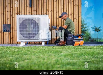 Ein Techniker wartet eine Klimaanlage, die sich neben einem modernen Haus befindet. In Arbeitskleidung verwendet er Werkzeuge aus einem organisierten Werkzeugkasten Stockfoto