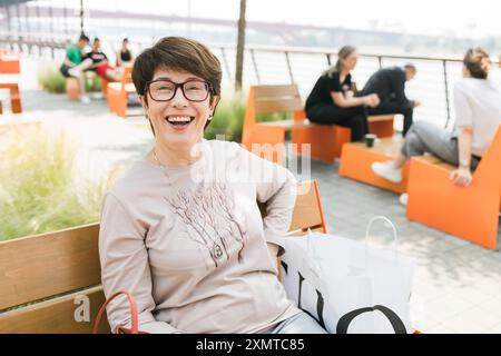 Außenporträt der schönen und eleganten lachenden mittleren Alter 55 - 60 Jahre alte Frau mit langärmligen Oberteil sitzend auf einer Bank auf der Straße. Spa kopieren Stockfoto