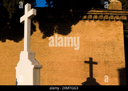 Weißes Kreuz, das während des Sonnenuntergangs am Cementerio de San Fernando in Sevilla, Andalusien, einen Schatten auf eine Ziegelmauer wirft. Stockfoto