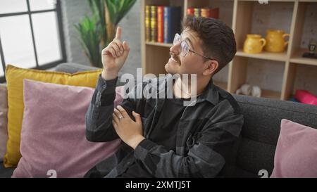 Ein nachdenklicher hispanischer Mann mit Schnurrbart sitzt bequem drinnen und zeigt nach oben, als hätte er eine Idee. Stockfoto