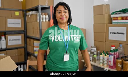 Lächelnde hispanische Frau, die im Lager steht, mit Spenden und Vorräten im Hintergrund. Stockfoto