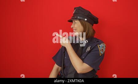 Eine lächelnde Polizistin zeigt auf etwas außerhalb der Kamera vor rotem Hintergrund. Stockfoto