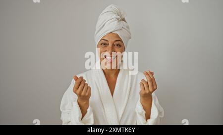 Ältere brünette hispanische Frau in Bademantel und Handtuch macht eine Geldgeste, während sie lächelt und vor einem isolierten weißen Hintergrund steht. Stockfoto