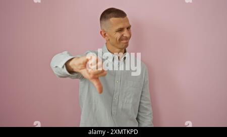 Junger Mann, der sich isoliert vor einem rosafarbenen Hintergrund mit den Daumen nach unten zeigt und mit einem beiläufigen Ausdruck und einer Geste Missbilligung zeigt. Stockfoto