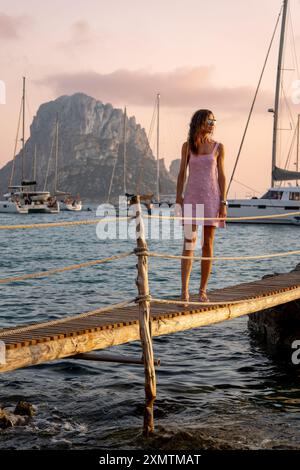 Junge Frau in pastellrosa Kleid bei Sonnenuntergang am Cala DHort Pier mit es Vedra Stockfoto