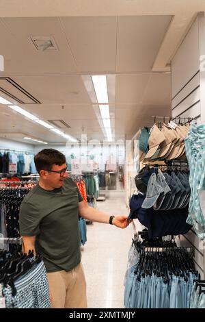 Ein Mann probiert einen blauen Hut an, während er eine Auswahl an Hüten in einem Bekleidungsgeschäft durchsucht. Männlicher Konsum, Second-Hand-Shopping Stockfoto