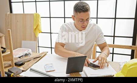 Ein junger hispanischer Mann arbeitet aufmerksam in einer Tischlerei mit einem Tablett und umgeben von Holzbearbeitungswerkzeugen. Stockfoto