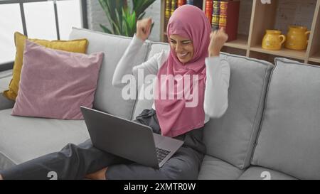 Eine aufgeregte Frau im Hijab feiert den Erfolg auf einem Laptop in ihrem modernen Wohnzimmer. Stockfoto