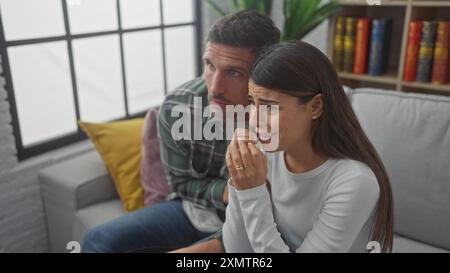 Ein hispanisches Paar, das ängstlich aussieht, während er zusammen auf einem Sofa im Wohnzimmer fernsieht. Stockfoto