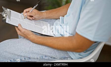 Hispanischer Mann in lässiger Kleidung sitzt drinnen und füllt Papiere auf einem Klemmbrett aus. Stockfoto